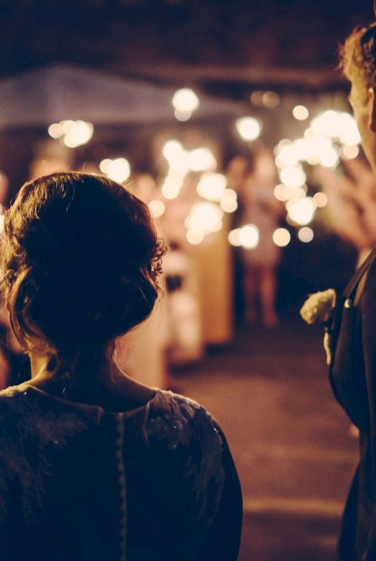 A couple stands among people holding sparklers, possibly celebrating an event or occasion in an outdoor setting at night.