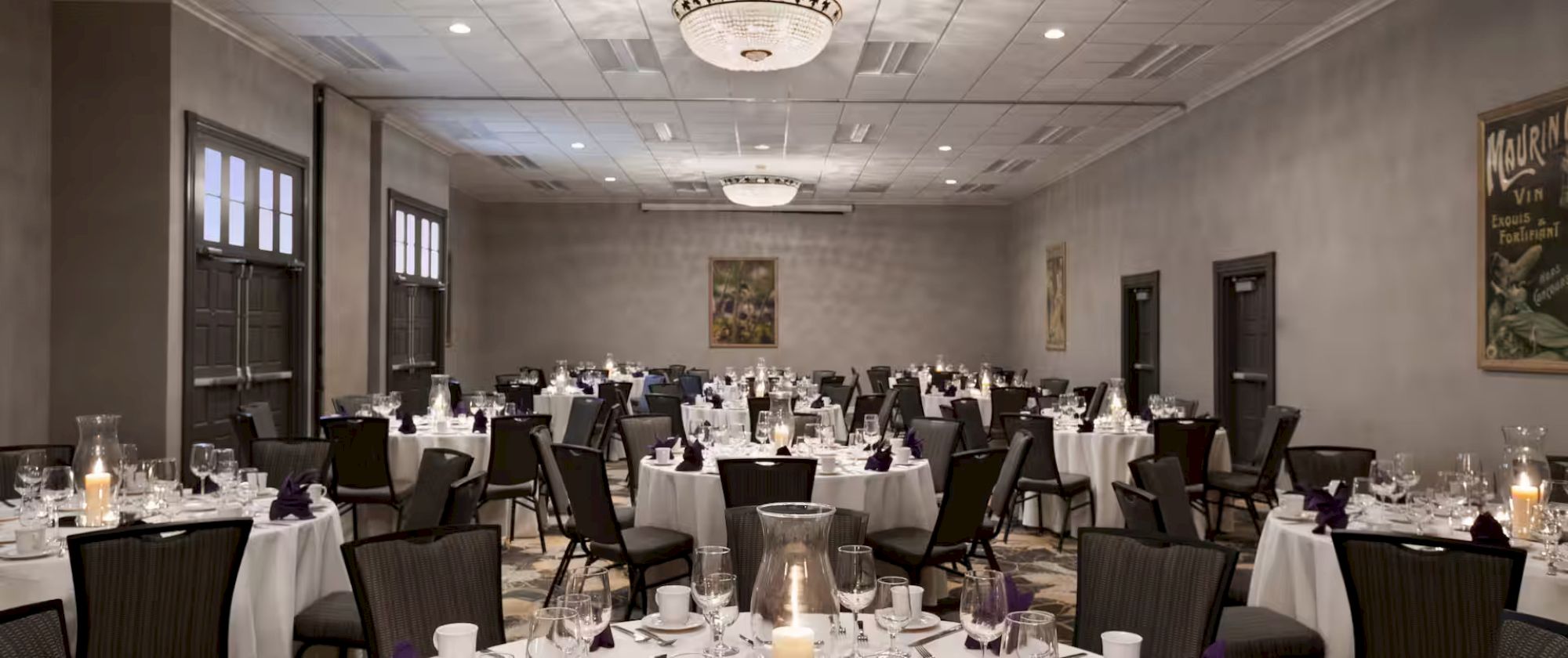 A banquet hall set for an event with round tables, white tablecloths, glassware, and candles, surrounded by patterned chairs.