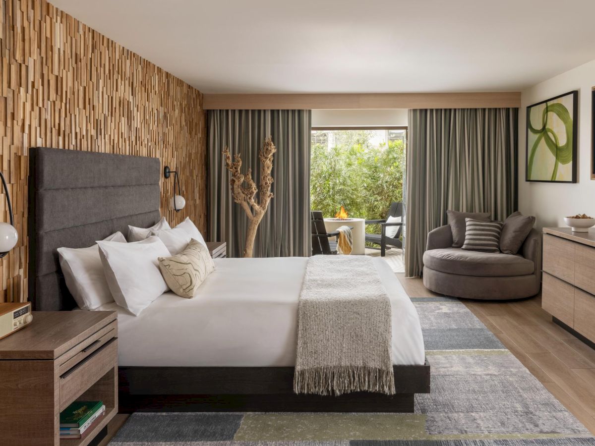 A modern hotel room with a large bed, geometric wood feature wall, nightstands, a seating area, and a balcony overlooking greenery, inviting relaxation.