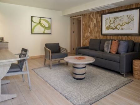 A modern living area with a gray sofa, two matching chairs, a round coffee table, a light rug, and a textured brown accent wall, plus wall art and a small desk to the left.