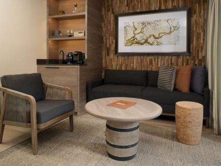 A cozy living nook with a dark sofa, wooden armchair, round marble coffee table, a textured wall, and a framed art piece, plus a cork stool.