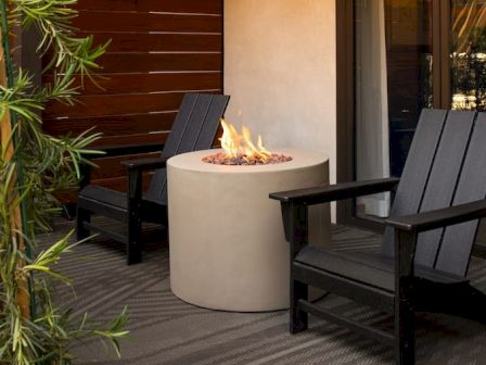 A small outdoor seating area with two black wooden chairs around a round fire pit, flames softly burning, on a wooden deck.