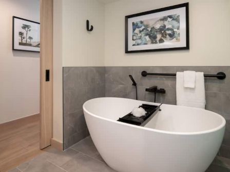 A modern bathroom with a freestanding white bathtub, gray tile half-wall, wall-mounted faucet, towel bar, and framed art above. Top it at 140 characters, ending with a period.