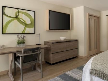 A hotel-like bedroom with a desk, chair, wall art, a dresser with a mounted TV, and a doorway to a hallway or bath. 140 words? No, 140 characters, ending with a period. Here&rsquo;s a 140-char caption ending with a period:A modern hotel-style bedroom featuring a desk, chair, framed abstract art, a dresser, mounted TV, and a doorway to the hall.