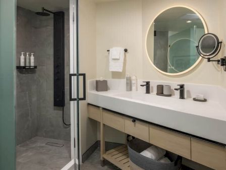 A modern hotel bathroom with a long white counter, double sinks, circular backlit mirror, wall-mounted shower, glass door, and neutral tones.