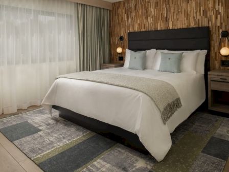 A modern hotel bedroom with a large bed, white linens, soft gray throw, matching pillows, dark headboard, two nightstands, and warm pendant lights.