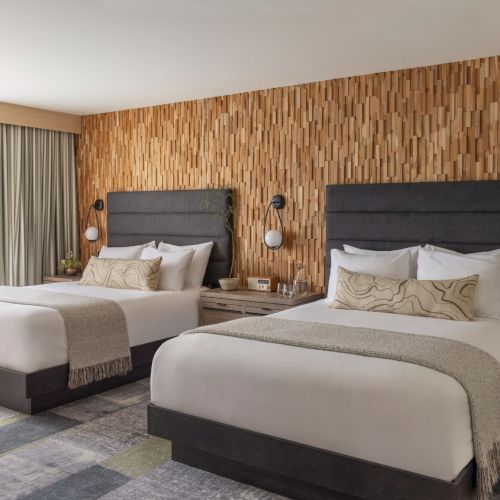 A modern hotel room with two queen beds, wooden feature wall, bedside lamps, and a balcony with curtains across from the beds.
