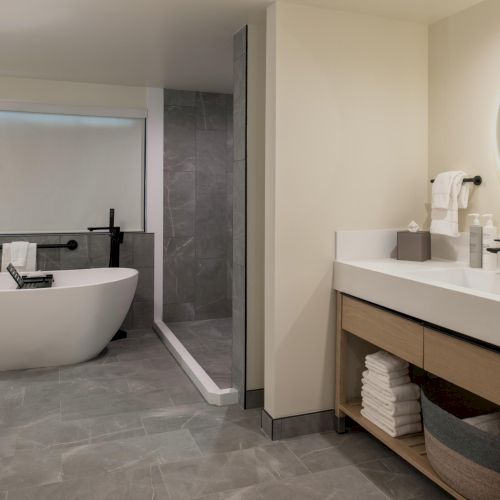 A modern bathroom with a freestanding tub, glass shower, and a wide vanity with round mirror and organized shelves.