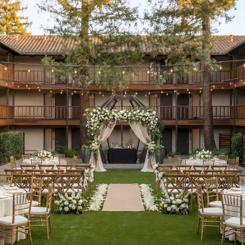 An outdoor wedding setup in a courtyard with a wooden arbor, floral arches, round tables with white linens, gold chairs, and string lights. Ending with a period.