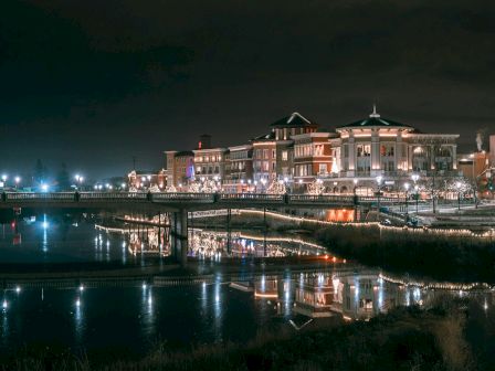 A nighttime riverside cityscape with lit buildings, a bridge, and reflections on calm water, glowing softly along the quay.